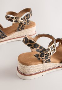 Sandales compensées imprimé léopard avec brides à la cheville, boucles dorées, semelles intérieures rembourrées couleur sable et semelles plateformes en jute tressé sur fond blanc.
