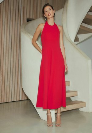 Femeie stând lângă o scară albă curbată, purtând o rochie midi roșie fără mâneci și pantofi cu barete bej la gleznă.