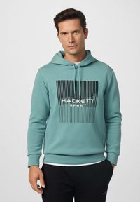 Sudadera color turquesa con un diseño gráfico de rayas negras y texto "HACKETT SPORT". Cuenta con cordones ajustables y puños acanalados.