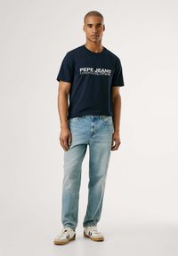 Dunkelblaues Baumwoll-T-Shirt mit weißem "PEPE JEANS" Logo, kombiniert mit hellblauen Jeans und weißen Sneakern mit schwarzen Akzenten.