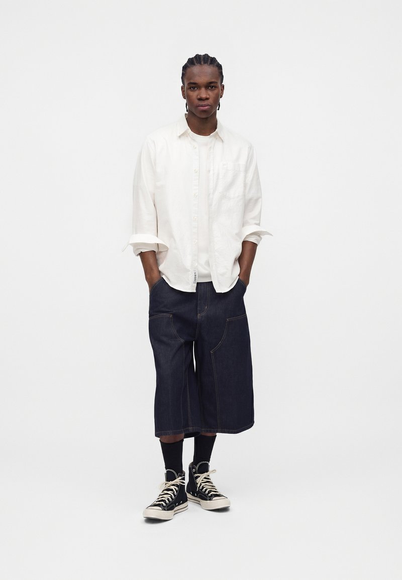 Ung mand med fletninger har en hvid skjorte med knapper, hvid undertrøje, mørke vidbens denimshorts, sorte sokker og sorte høje sneakers.