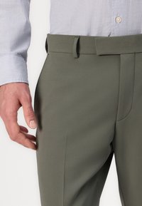 Sötétzöld nadrág sima, szabott anyagból, lapos elejű kialakítással. A derékpánt kampós és bársonyos záródással rendelkezik, látható zsebek nélkül.