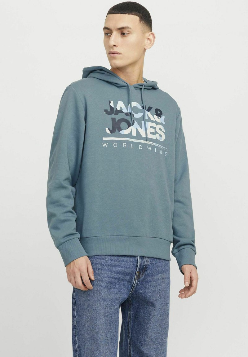 Sudadera con capucha en color azul verdoso con un estampado de logo "JACK & JONES" en blanco y azul, con puños y dobladillo acanalados, combinada con jeans azules.