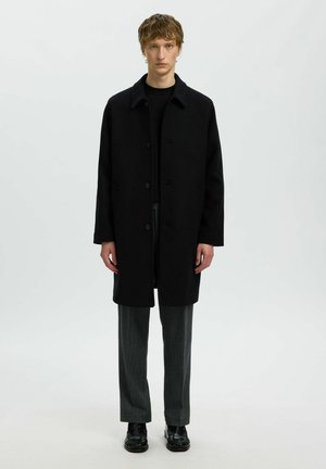 Jeune homme portant un manteau noir boutonné, un pantalon gris foncé et des chaussures noires, debout devant un fond blanc uni.