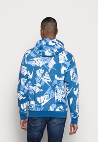 Modrá mikina s kapucí s bílými logy Nike, vyrobená z měkké látky, vybavená kapucí, žebrovanými manžetami a lemem, s volnějším střihem.