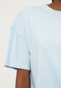 Φωτεινό γαλάζιο βαμβακερό κοντομάνικο μπλουζάκι που φοριέται από άτομο με μεσαίο προς σκούρο τόνο δέρματος, εμφανιζόμενο από τον ώμο έως το μέσο του κορμού.