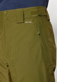 Olive green pants with a smooth texture, featuring a button closure, Velcro accents, and a zippered side pocket; brand logo visible.