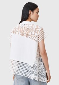 Top blanco de manga corta con detalles de encaje con patrones florales y un diseño de rejilla, que contrasta con un panel trasero blanco sólido.
