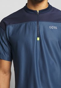 Námořnická sportovní košile s krátkým rukávem a zipem, vysokým límcem a strukturovanou tkaninou, s jemným logem na hrudi. Neonový akcent poblíž zipu.
