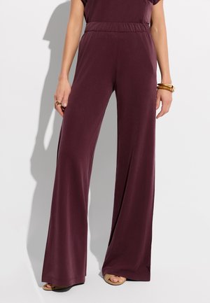 Kobieta nosząca wysokie, szerokie ciemnofioletowe spodnie z elastycznym pasem, brązowe sandały z odkrytymi palcami oraz złote bransoletki na prawym nadgarstku.