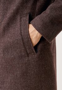 Tessuto marrone con texture e un motivo sottile, dotato di una tasca laterale e una mano visibile all'interno. La manica è leggermente arrotolata.