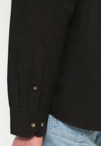 Manga larga de camisa marrón oscuro con dos botones, llevada sobre el bolsillo de unos jeans de denim azul claro.