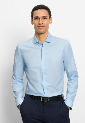 Mann trägt hellblaues Hemd mit Knöpfen und marineblaue Hose mit schwarzem Gürtel, steht mit den Händen in den Taschen vor weißem Hintergrund.