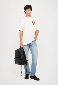 White oversized t-shirt with red heart graphic, light blue jeans, black backpack, and black shoes. Plain background highlights details.