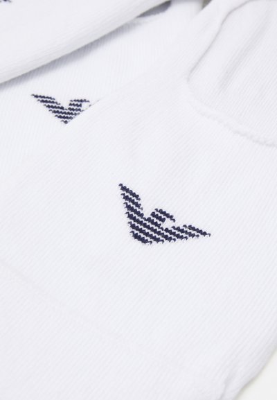 Chaussettes blanches à texture côtelée, avec un logo brodé bleu marine en forme d'aile stylisée. La vue rapprochée met en avant les détails du design.