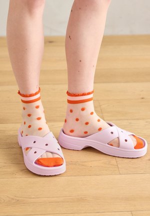 Jambes portant des chaussettes blanches transparentes à pois orange et à volants en haut, associées à des sandales légères violet clair sans bride sur un sol en bois.