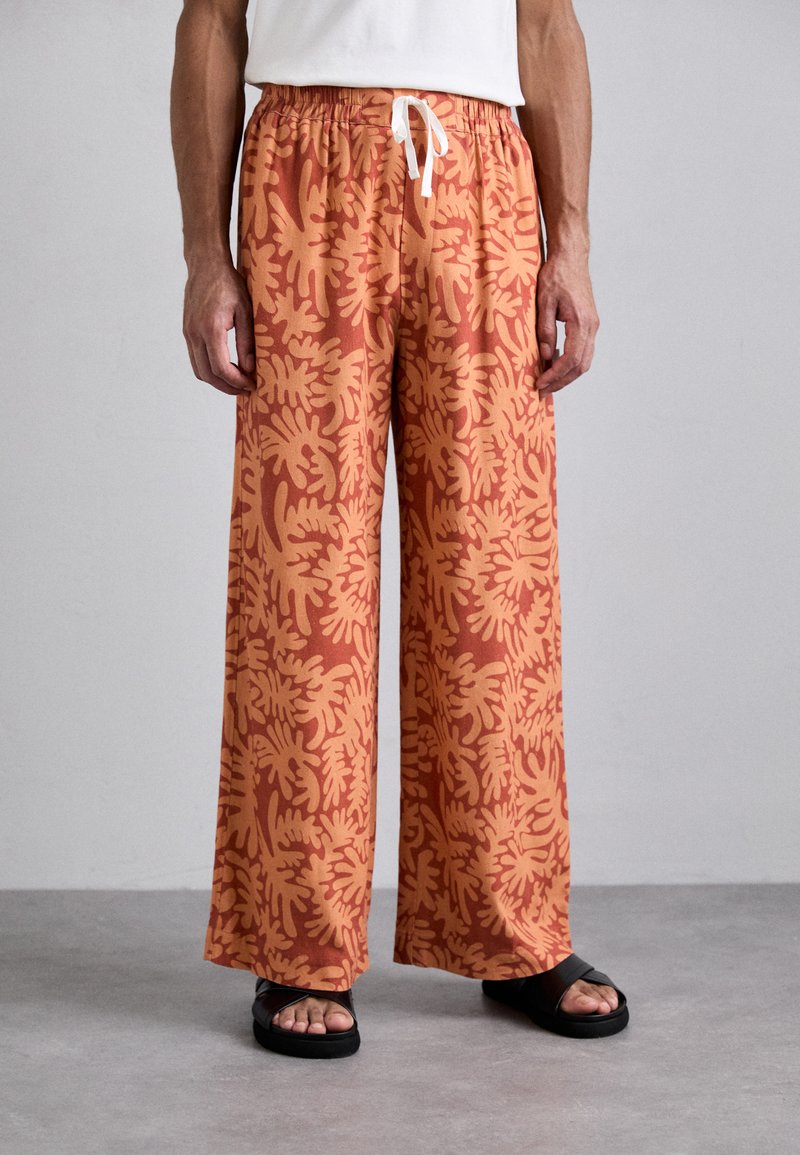 Pantalons à jambe large en orange brûlé avec un grand motif de feuille corail. Dotés d'une taille élastique avec un cordon blanc. Tissu texturé.
