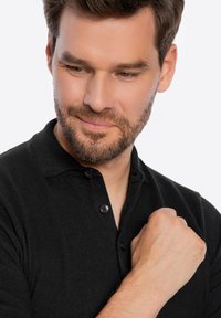 Schwarzes Polohemd aus strukturiertem Stoff, mit zwei Knöpfen am Kragen und gerippten Details. Das Modell hat einen leichten Bart und schaut nach unten.