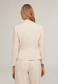 Blazer ajusté de couleur beige clair avec des manches longues, des épaules structurées et de subtiles coutures diagonales. Texture lisse, détails minimalistes et sans quincaillerie.