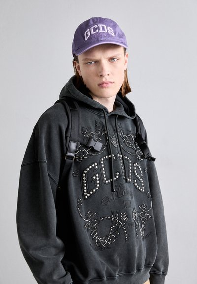 Felpa nera con design di borchie e logo sul davanti, abbinata a un cappellino viola con testo ricamato. Le cinghie dello zaino sono visibili sulla spalla.