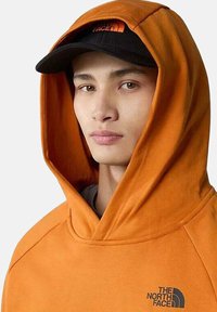Sudadera con capucha naranja y un logo negro, con una textura suave. Se lleva una gorra negra con un logo naranja, enmarcando el rostro.