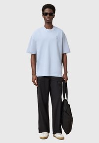 Lichtblauw oversized T-shirt met korte mouwen, klein logo op de borst, gecombineerd met zwarte oversized broek en witte sneakers, met een zwarte tas in de hand.