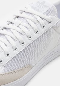 Bílá teniska z materiálů síťoviny a semiše, s texturovaným povrchem, bílými tkaničkami a jazykem s logem značky. Řady dírek na tkaničky.