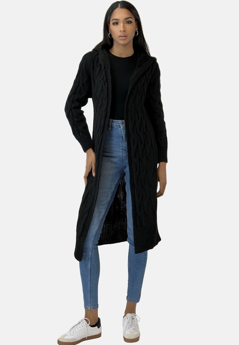 Langer schwarzer Strickcardigan mit Kapuze, der ein Zopfmuster und einen offenen Vordereingang hat. Getragen über einem schwarzen Oberteil und blauen Jeans, kombiniert mit weißen Sneakers.