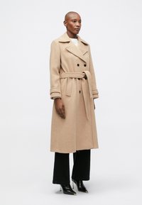 Beige gyapjúszövet trench coat, dupla gombolású dizájnnal, övvel a deréknál és hajtott gallérral. Fekete, bő szárú nadrággal és magassarkú csizmával párosítva.