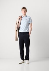 Tricou polo bleu deschis cu margini negre, sacou bej purtat pe umăr, pantaloni navy și adidași albi. Design simplu, fără modele notabile.