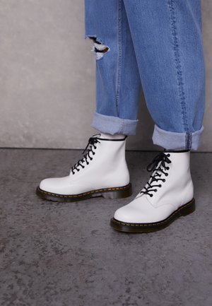 Bottes blanches en cuir avec des lacets noirs, des coutures jaunes et une semelle épaisse et transparente. Portées avec un jean bleu clair retroussé présentant des détails déchirés.