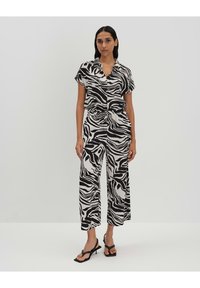 Une combinaison zèbre noir et blanc avec des manches courtes, une taille cintrée et des pantalons larges. Portée avec des sandales plates noires.