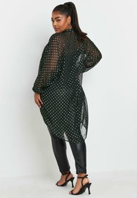 Copertura nera trasparente con pois bianchi, maniche lunghe e design morbido. Indossata sopra pantaloni neri aderenti e sandali con tacco.