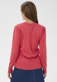 Pull tricoté corail à manches longues avec ourlet côtelé. Présente un détail froncé à l'épaule et une texture lisse et douce. Porté sur une jupe en jean.