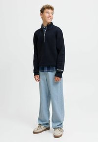 Donkerblauwe pullover sweatshirt met een hoge kraag en een halve rits, gecombineerd met een lichtblauw geruit overhemd eronder, en losse lichtgewassen spijkerbroeken.