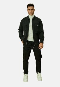 Veste zippée noire avec deux poches avant, associée à un pantalon cargo noir et des baskets blanches. Le col roulé en dessous est gris clair.