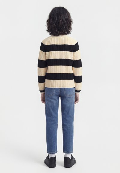 Παιδί με σγουρά μαλλιά που φοράει πουλόβερ με κρεμ και μαύρες ρίγες, μπλε τζιν, λευκές κάλτσες και μαύρα παπούτσια, στέκεται με την πλάτη προς τα επάνω.