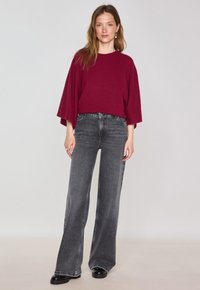 Pull en maille oversize bordeaux avec des manches larges, associé à un jean flare gris taille haute et des bottes noires chunky. Fond neutre.