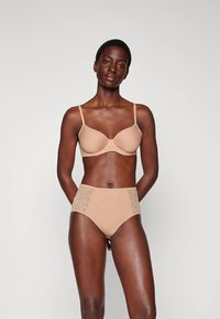 Smėlio spalvos liemenėlė su paminkštintomis kaušeliais ir reguliuojamomis petnešėlėmis, derinamas su aukšto liemens kelnaitėmis, kurių šonuose yra nėrinių panelės. Lygi tekstūra, be siūlių dizainas.