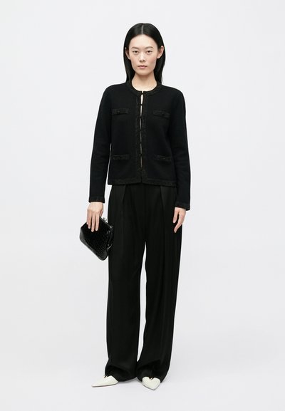 Γυναίκα που φοράει μαύρο μπουφάν και παντελόνι φαρδύ, κρατώντας μια μαύρη τσάντα με υφή, στέκεται μπροστά σε λευκό, απλό φόντο.