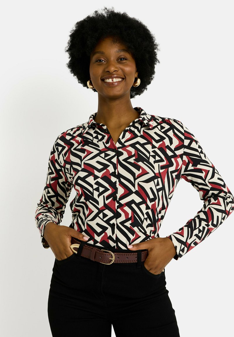 Chemise à manches longues présentant un motif géométrique noir, rouge et crème. Design à boutons avec col, portée avec une ceinture marron.