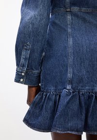 Vestido de ganga com mangas longas, bainha com volantes e botões decorativos. Apresenta uma cor azul desbotada e um tecido texturizado.