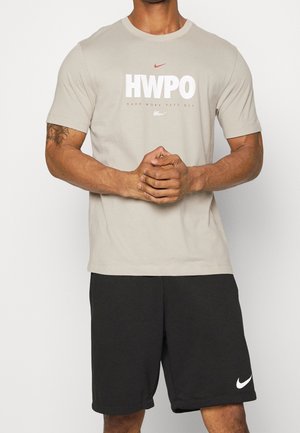 Man draagt een beige T-shirt met de tekst "HWPO Hard Work Pays Off" en zwarte Nike-shorts, staat met gevouwen handen voor zich op een effen achtergrond.