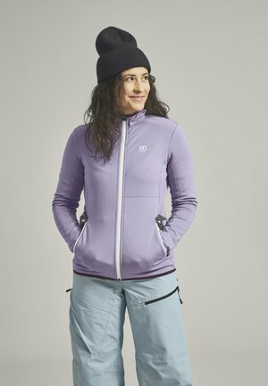 Purple zip-up jacket with side pockets, white zippers, and a high collar. Worn over light blue pants and paired with a black beanie.