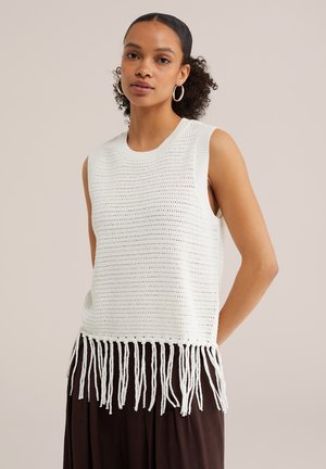 Vrouw met krullend haar draagt een wit mouwloos gebreid topje met franjes aan de zoom en een bruine broek tegen een effen beige achtergrond.