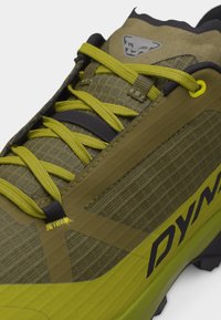 Arčiau alyvuogių žalios ir geltonos spalvos pėsčiųjų batų su raštuota medžiaga, geltonomis batų raištelėmis ir juodai baltu gyvūno logotipu viršuje.