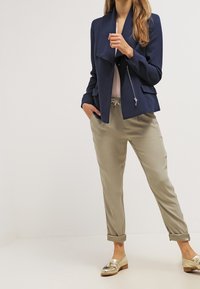 Námořnicky modrá bunda s volně splývajícím límcem a kapsami na zip, doplněná světle béžovými kalhotami a metalicky zlatými botami. Hladké textury tkaniny.