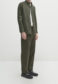 Camicia verde oliva con bottoni e colletto, abbinata a pantaloni verde scuro e scarpe nere. Tessuto morbido e vestibilità rilassata.