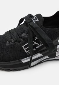Scarpa sportiva nera con tomaia in maglia, lacci piatti e suola ammortizzata, con dettagli argento e logo sul lato.