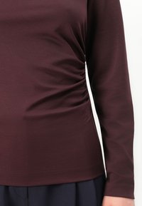 Close-up van een persoon die een lange mouw, gerimpelde donker bordeaux top draagt met marineblauwe broek, waarbij de textuur van de stof en het detail van de mouw zichtbaar zijn.